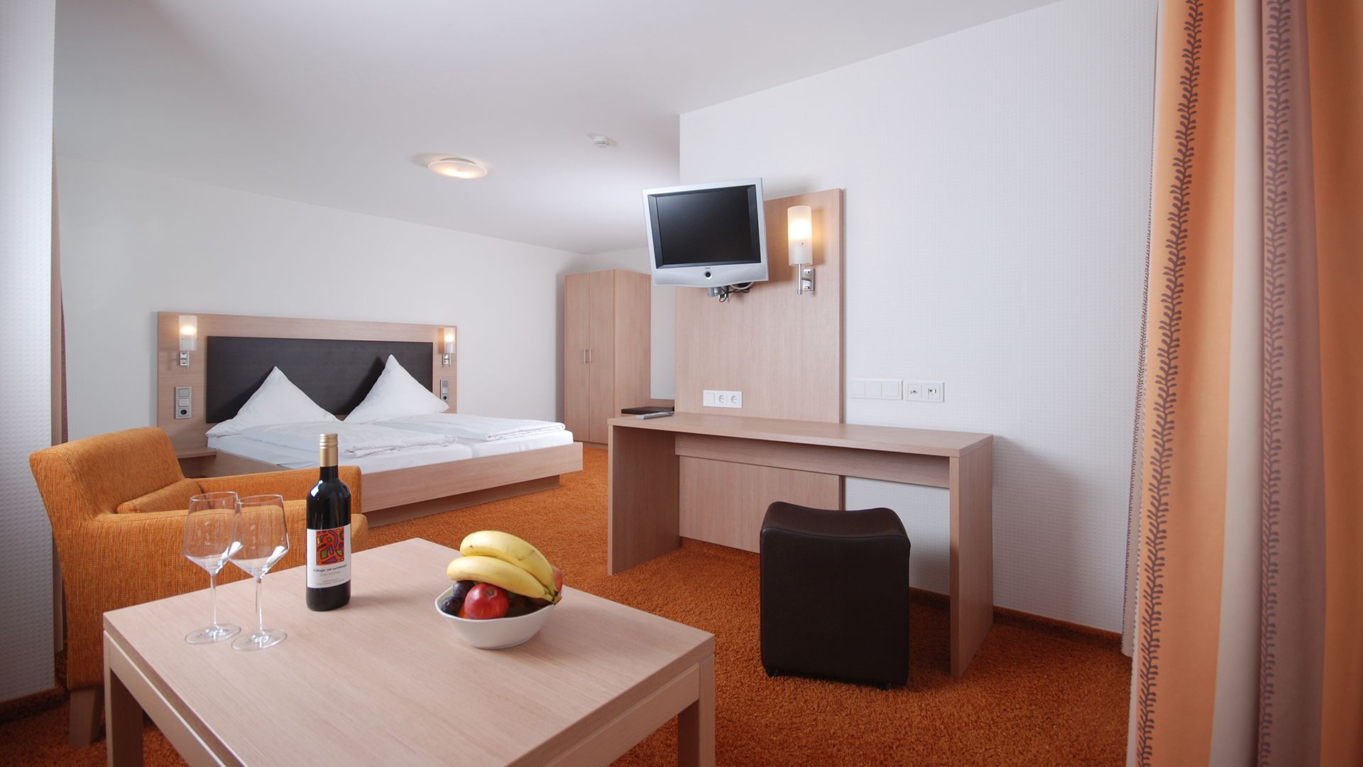 Habitación de hotel con cama, escritorio, silla, televisión y una pequeña mesa con una botella y vasos.