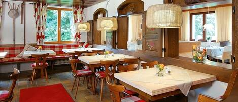 A restaurant with wooden tables and chairs, a red and white checkered bench, and large windows with curtains.