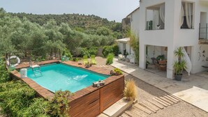 Zona de piscina moderna amb un solàrium de fusta, envoltada de vegetació i un edifici blanc amb grans finestres.