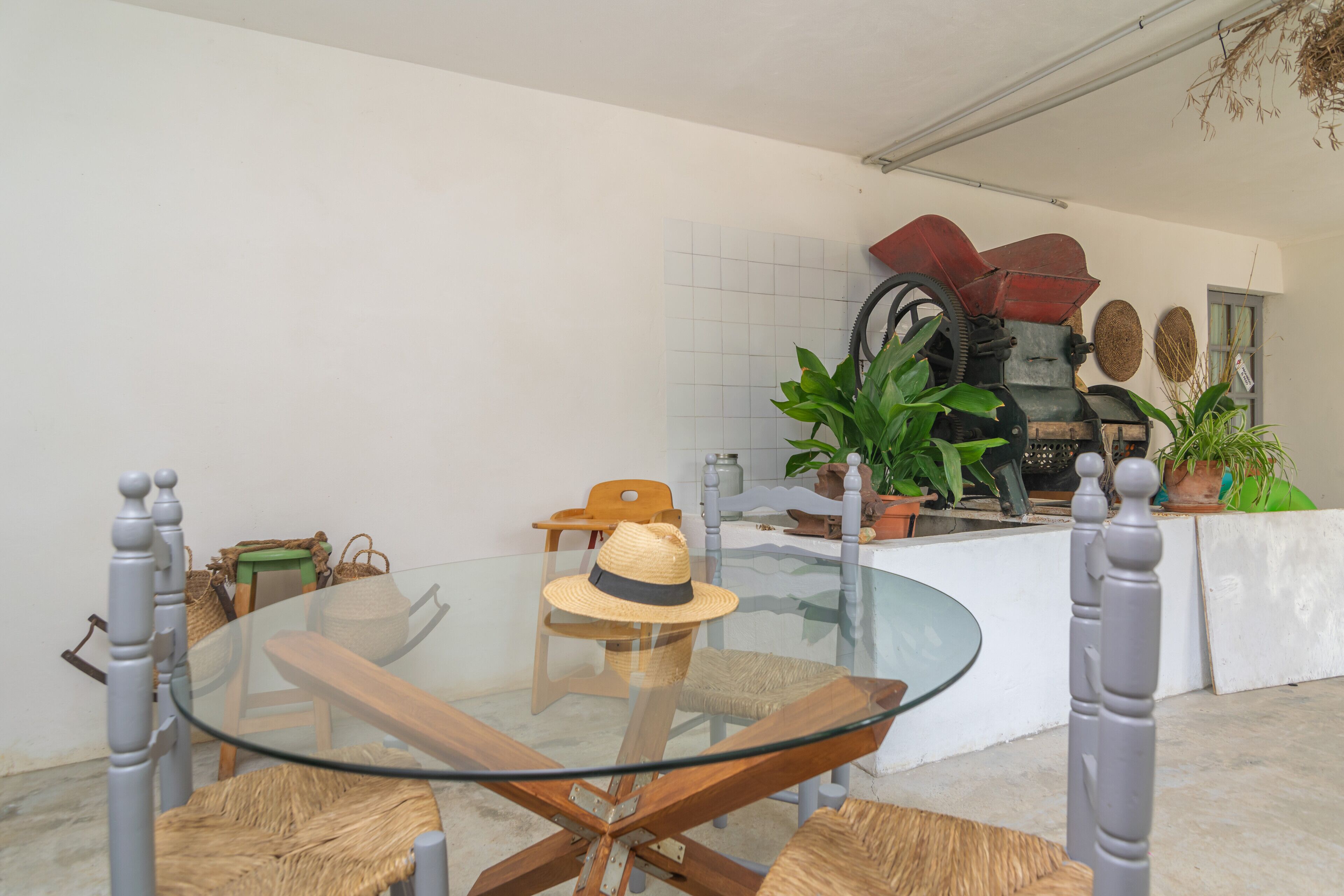 A dining area with a glass table and wooden chairs, a wicker chair with a hat, and a rustic outdoor grill.