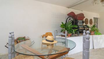 A dining area with a glass table and wooden chairs, a wicker chair with a hat, and a rustic outdoor grill.