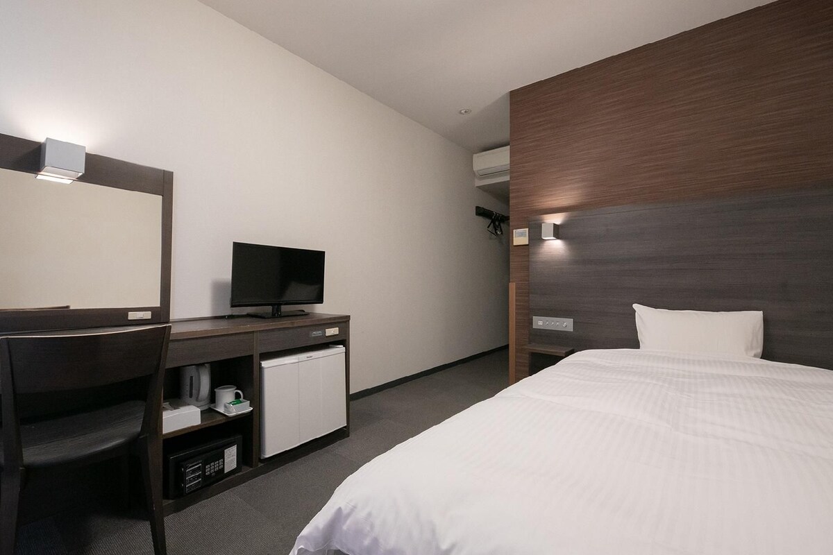 A hotel room with a bed, a desk with a television, a chair, a small cabinet, and a wall-mounted air conditioning unit.