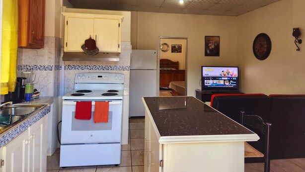 A compact kitchen with a stove, oven, and refrigerator. There is a dining area with a table and chairs. A television is mounted on the wall. - Stewart Suite Apt -Trincity, Airport, Full Apt (Trincity)