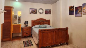 A bedroom with a wooden bed, a nightstand, a dresser, and a chandelier. There are three wall-mounted paintings and a mirror. - Stewart Suite Apt -Trincity, Airport, Full Apt (Trincity)