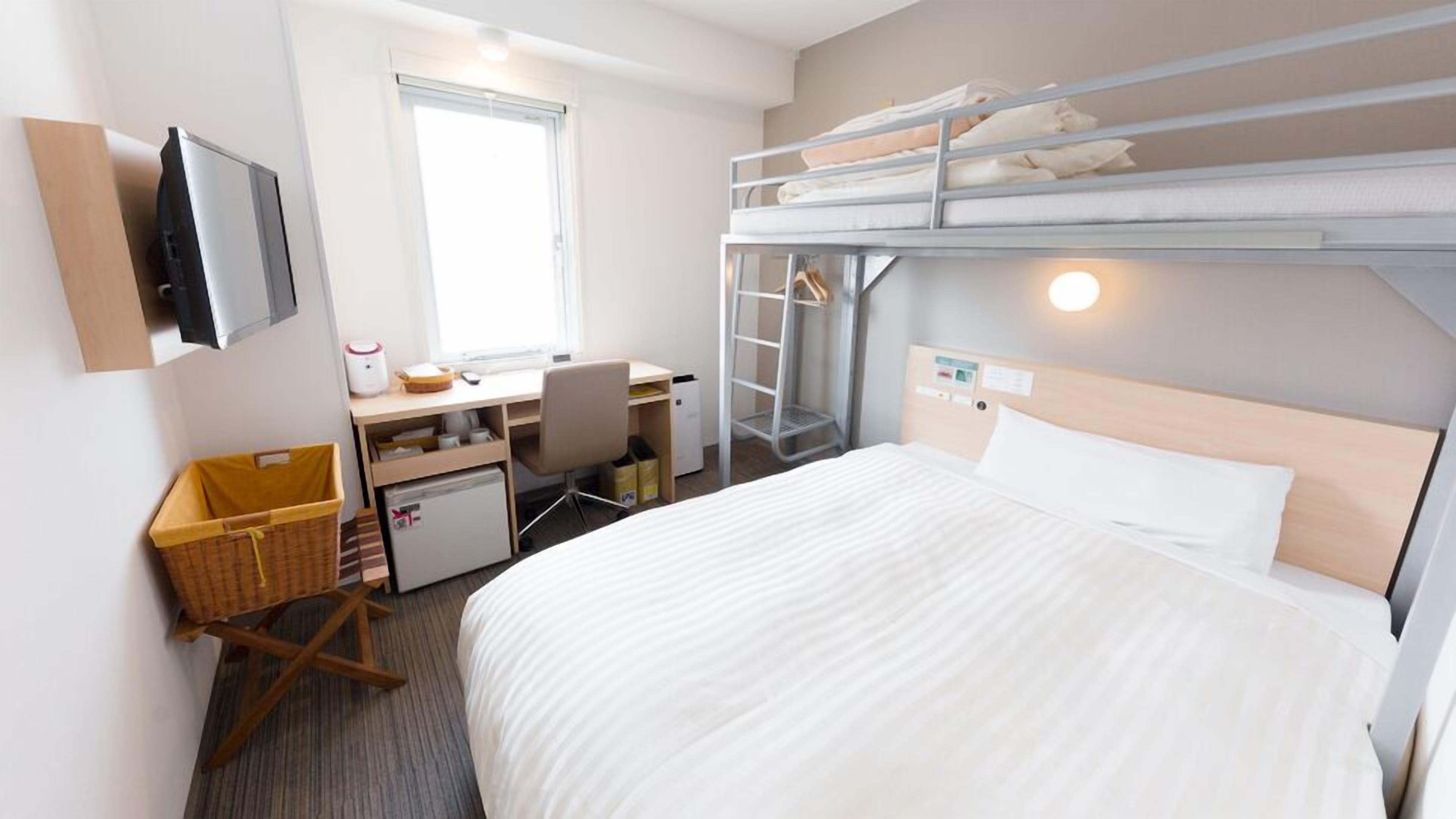 A hotel room with a bunk bed, a desk with a chair, a small refrigerator, a TV, and a window.