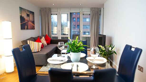 A modern living room with a sofa, dining table, chairs, and a TV. - The Pinnacle by Jim Paul Apartments (Reading)