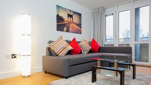 A modern living room with a grey sofa, colorful striped pillows, a glass coffee table, and a large window with curtains. - The Pinnacle by Jim Paul Apartments (Reading)
