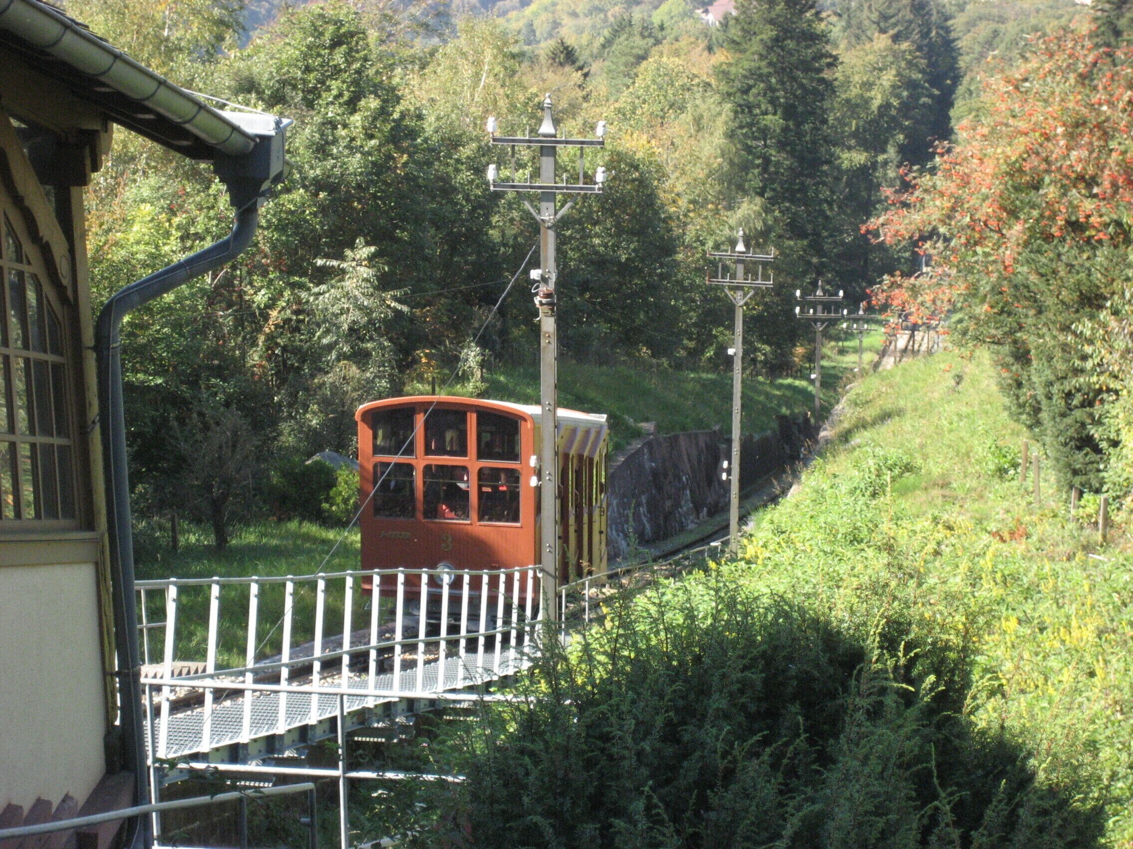 Sistema de teleférico com um carro vermelho sobre trilhos, cercado por vegetação exuberante e uma encosta.
