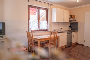 A compact kitchen with wooden cabinets, a dining table with chairs, a window with curtains, and a built-in dishwasher.