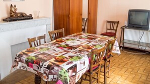 A dining area with a table covered in a coffee-themed tablecloth, wooden chairs, a wooden wardrobe, a television on a stand, and a marble countertop.