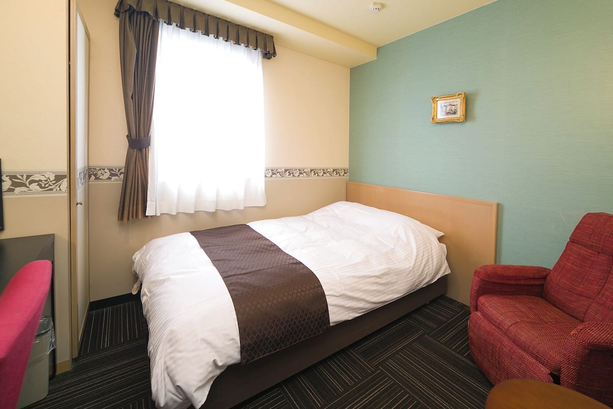 A hotel room with a bed, a red armchair, a television, and a window with curtains.