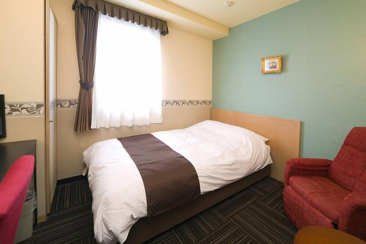 A hotel room with a bed, a red armchair, a television, and a window with curtains.