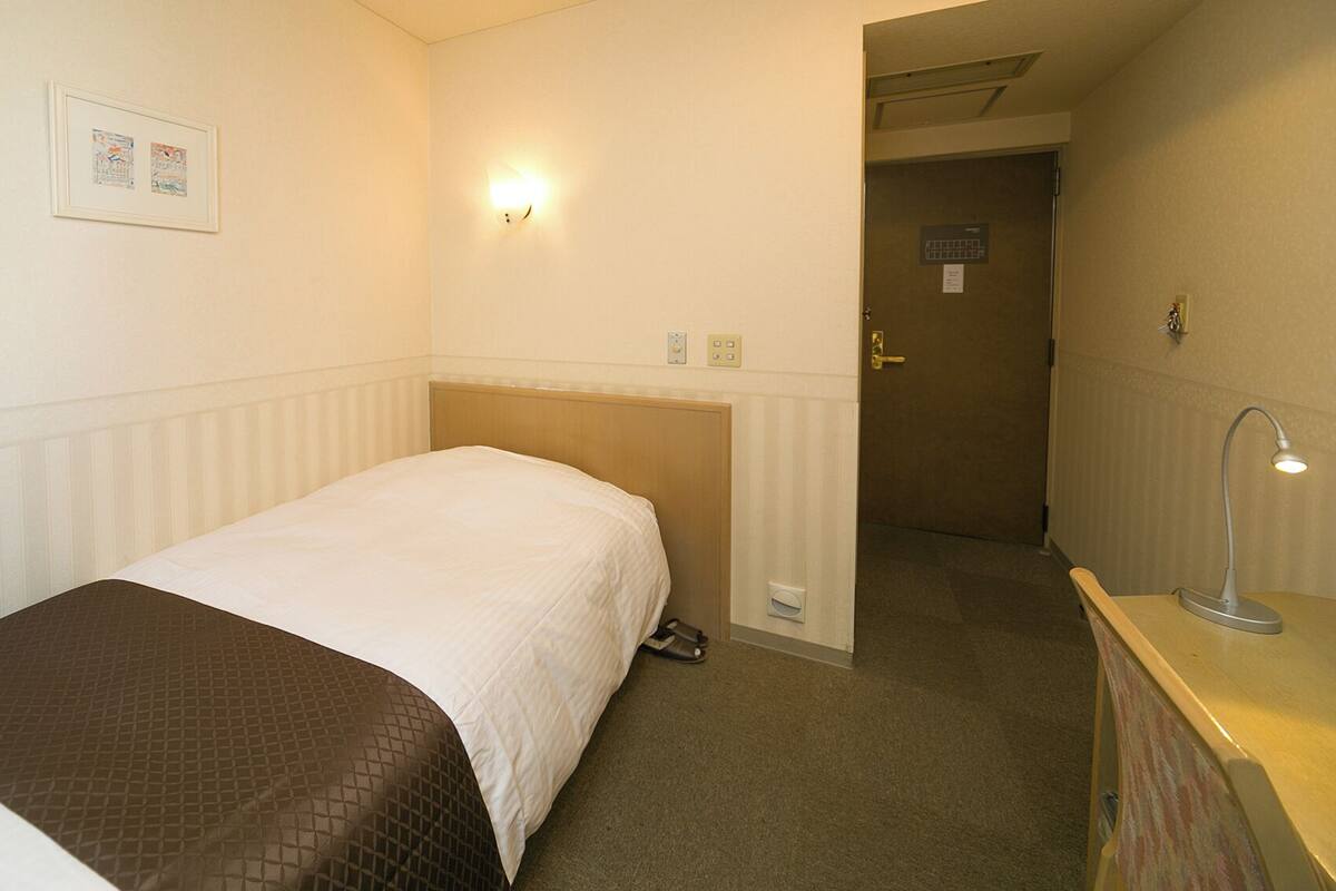 A hotel room with a bed, a desk with a lamp, a door, and a wall-mounted light fixture.