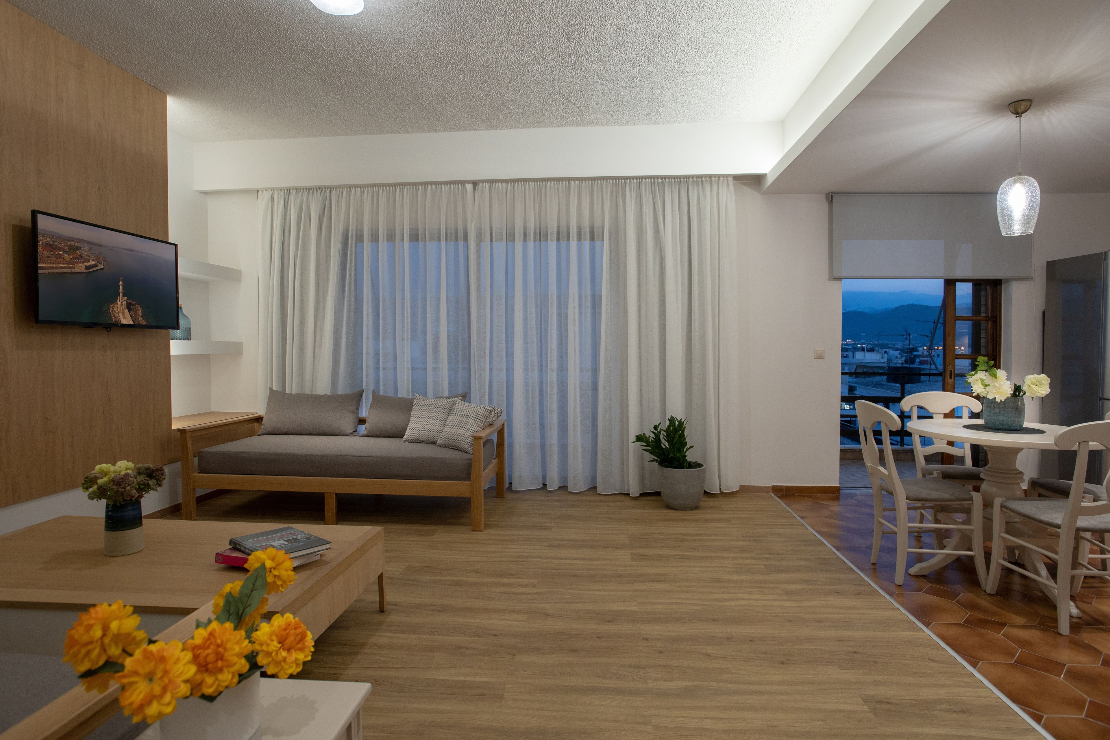 Un salon moderne comprenant un canapé, une table à manger avec des chaises, un téléviseur fixé au mur et une grande fenêtre avec des rideaux.