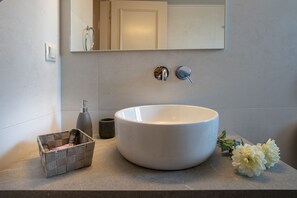 Une salle de bain avec un grand lavabo blanc, un miroir, un petit panier en osier, un récipient foncé et deux fleurs blanches posées sur le comptoir.