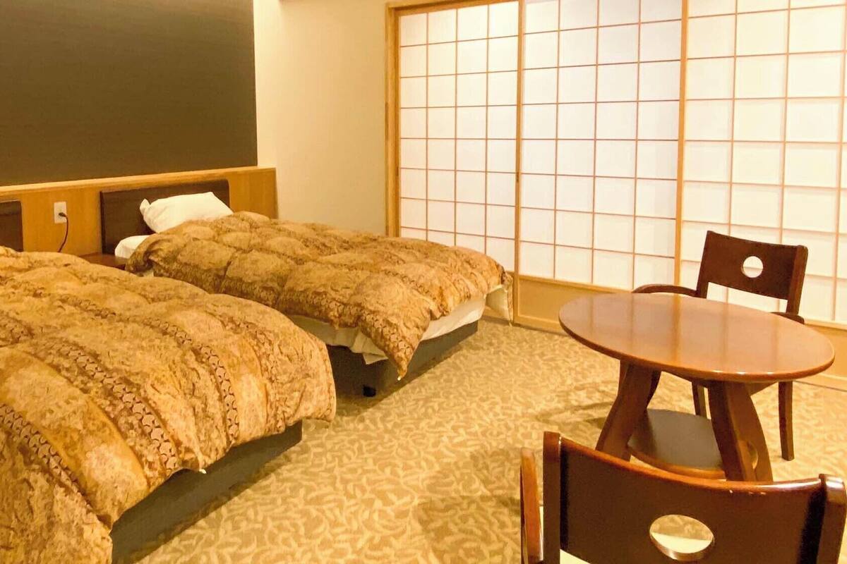 Habitación de hotel con dos camas, cabecero de madera, pared con esteras de tatami, una mesa redonda de madera y una silla.