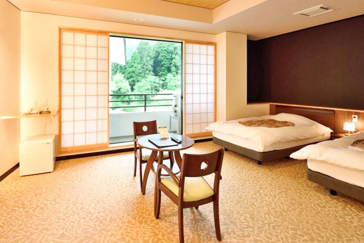 Una habitación de hotel con una estética japonesa tradicional, que presenta suelo de tatami, una mesa baja con sillas, una cama con cabecera y un ventanal corredero que da a un balcón.
