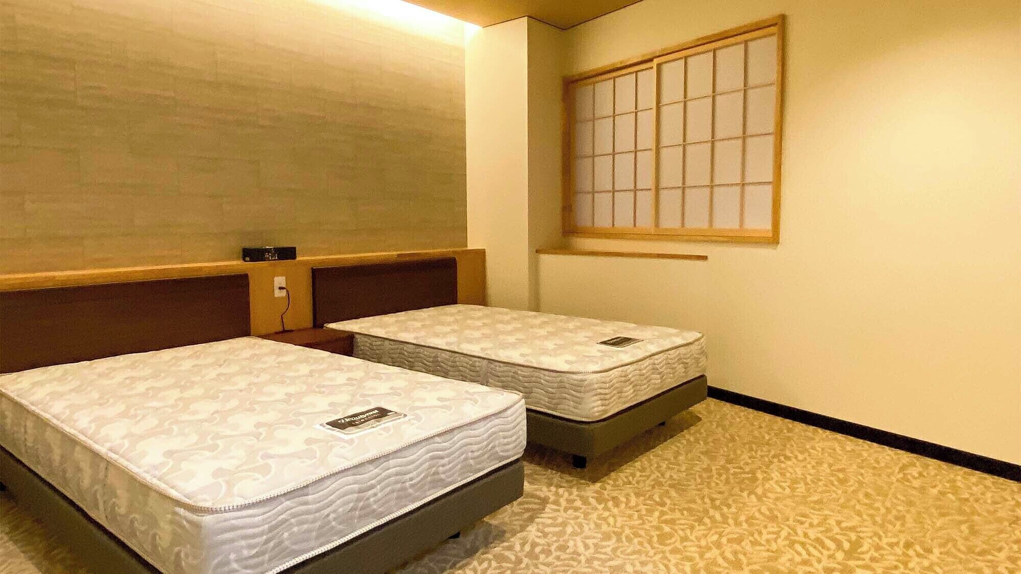 A room with two beds, a window with a grid pattern, and a wall-mounted electrical outlet.