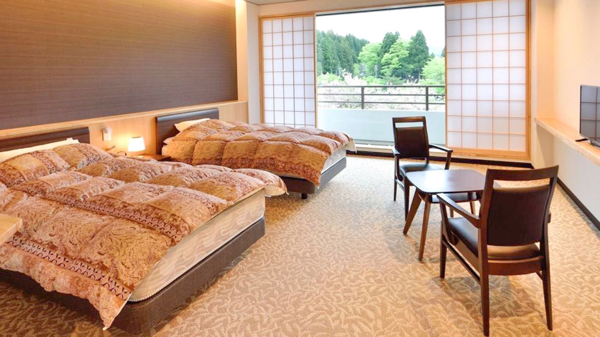 A hotel room with two beds, a sitting area with a table and chairs, and a view of a forest through large windows.