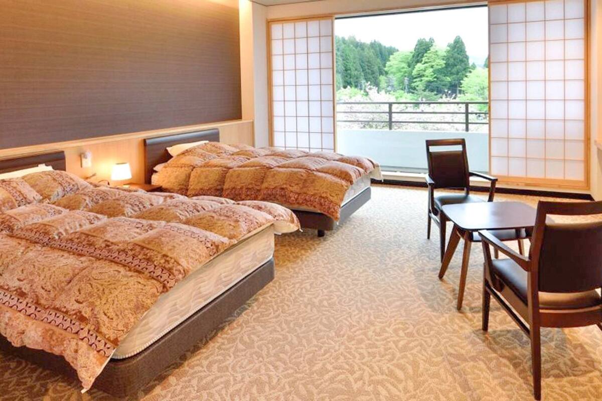 Habitación de hotel con dos camas, un espacio de descanso con una mesa y sillas, y vista a un bosque a través de amplios ventanales.