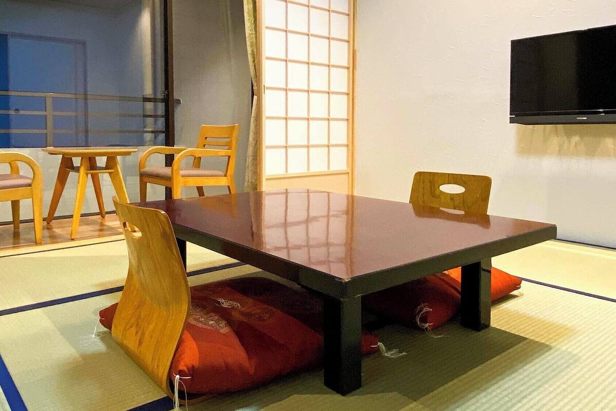 Una habitación japonesa tradicional con suelo de tatami, una mesa baja, sillas de madera y un televisor.