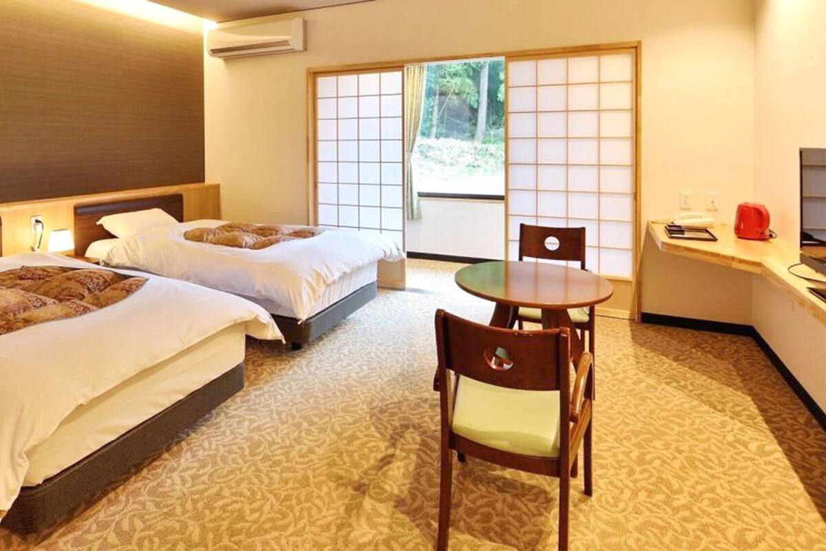 Habitación de hotel con dos camas, una mesita, una silla y vistas al exterior a través de una puerta corrediza.