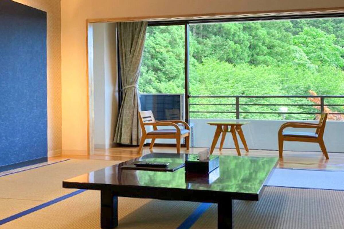 Una habitación con un ventanal grande, una televisión de pantalla plana, una mesa de madera y dos sillas de madera.