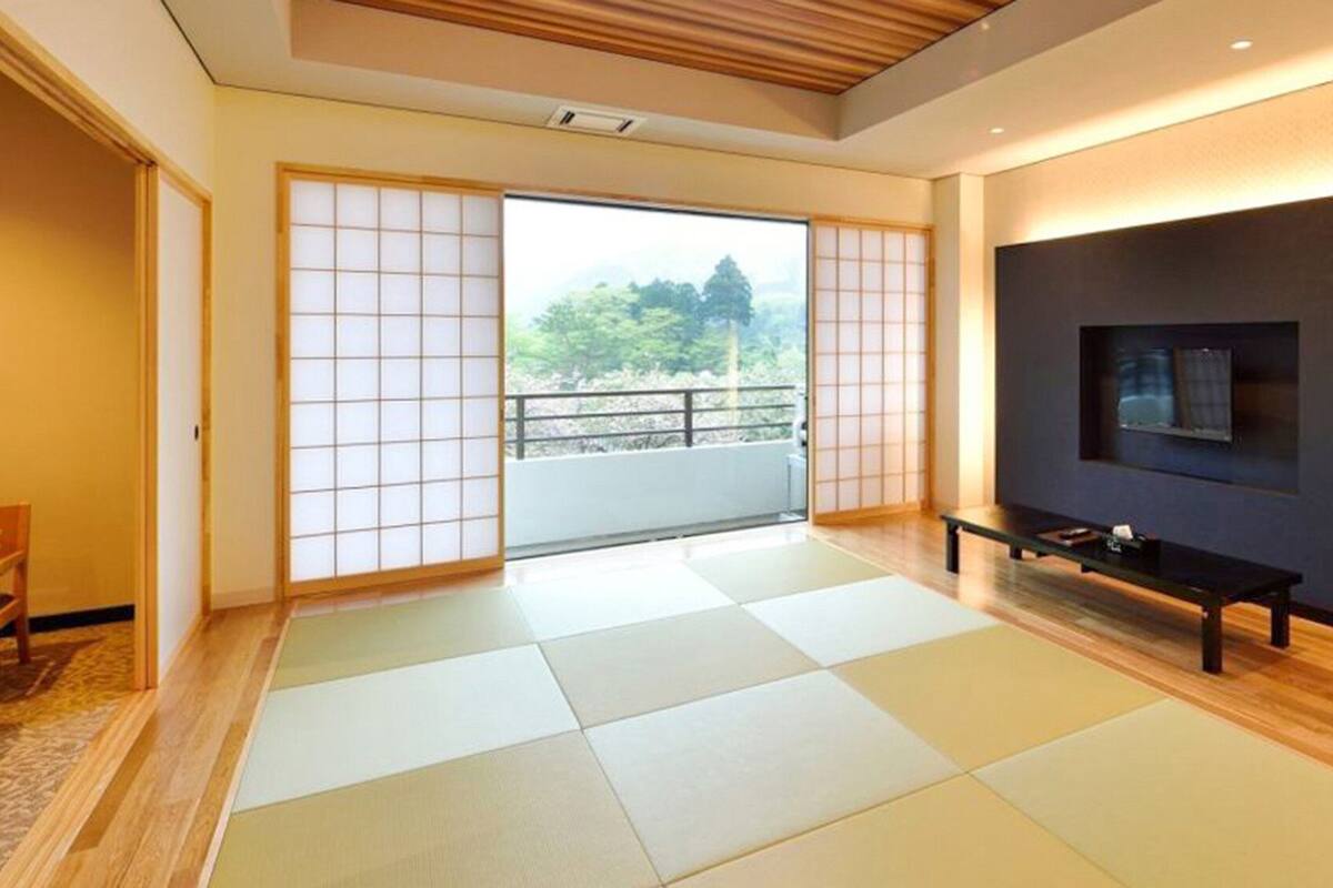 Una habitación con esteras de tatami, una puerta corredera hacia un balcón, un televisor montado en la pared y un banco.