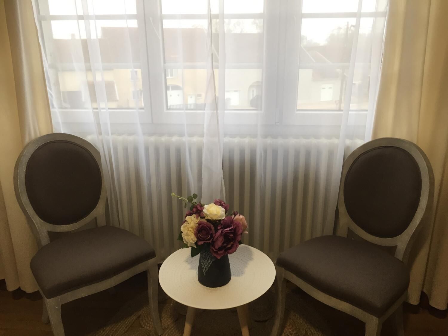 Two chairs facing each other with a small round table in between, a vase with flowers on the table, and a view of a building through large windows with sheer curtains.
