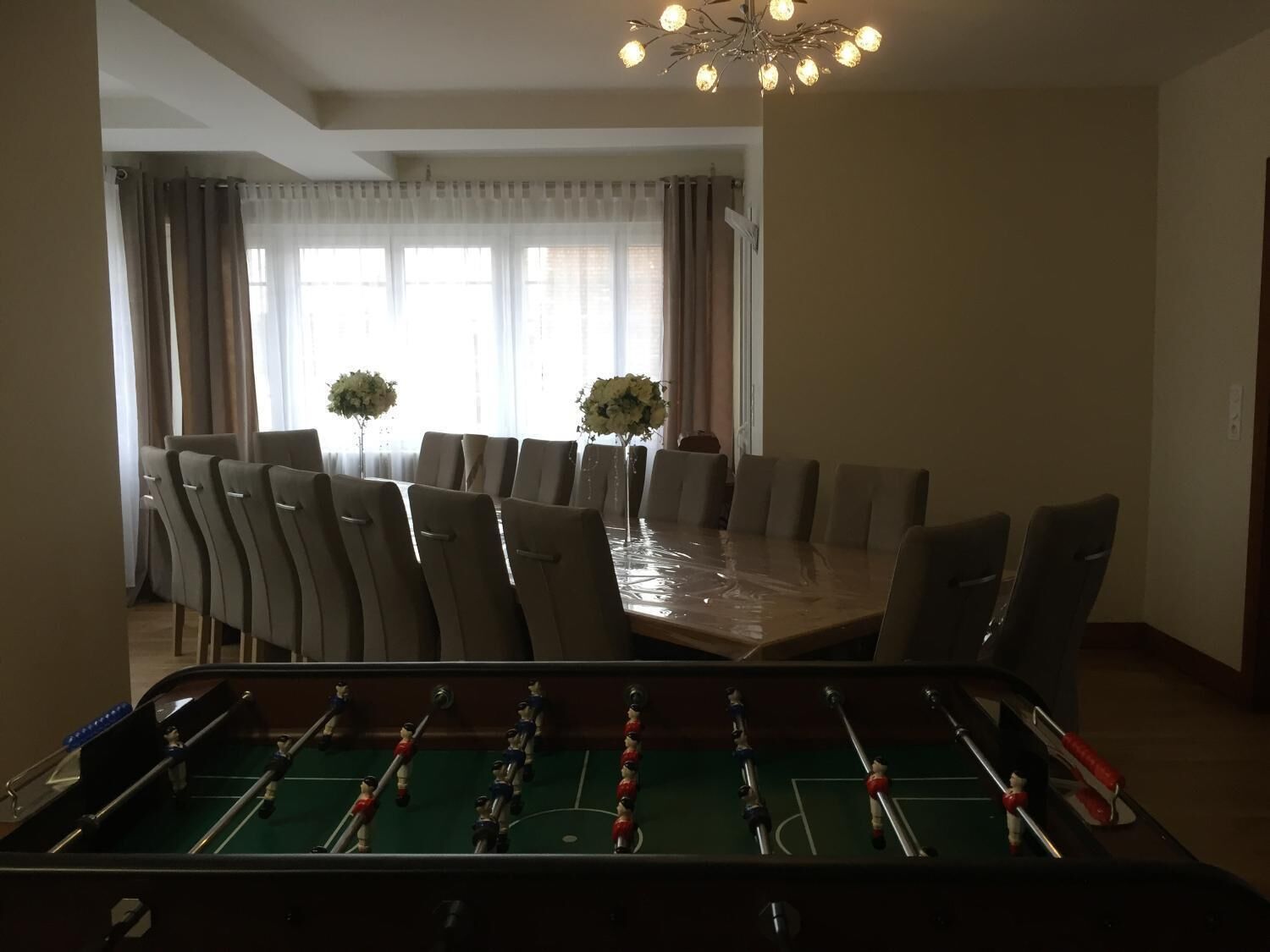 Un área de comedor con mesa de fútbol de mesa en primer plano, una mesa grande con sillas y una araña.