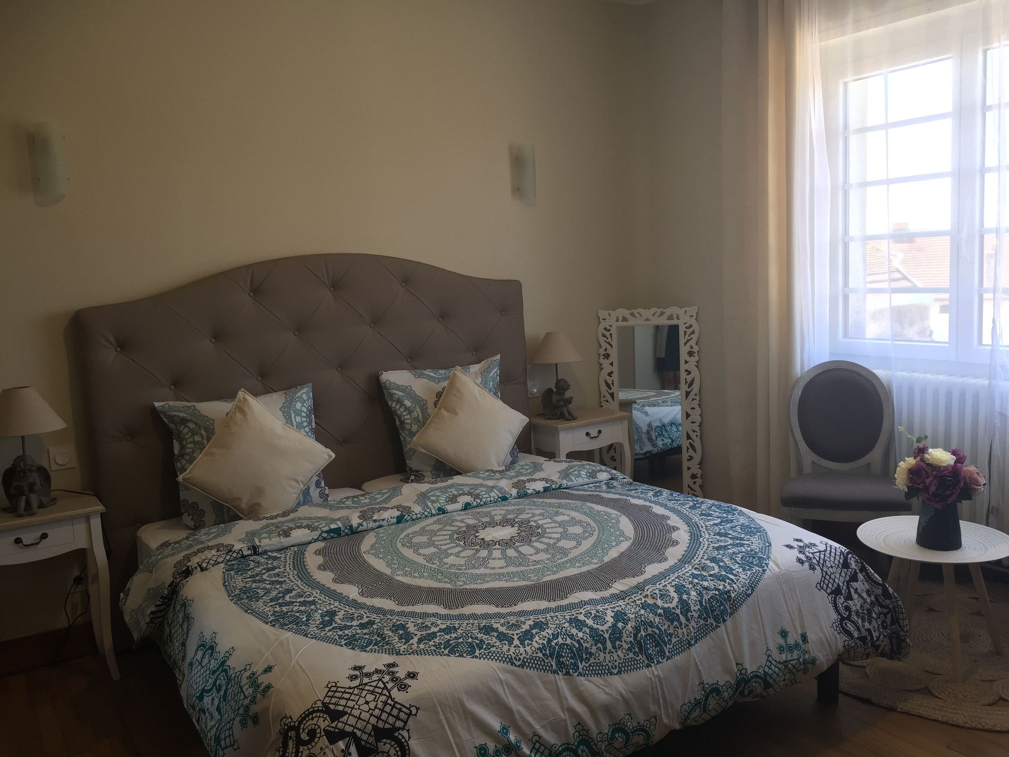 Een slaapkamer met een gestapeld hoofdbord, een groot bed met beddengoed met een patroon, nachtkastjes met lampen, een spiegel en een raam met gordijnen.