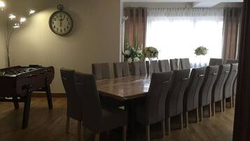 Et spiseområde med et langt træbord og flere stole, et ur på væggen og et vindue med gardiner.