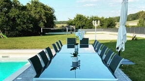 Une table de jardin rectangulaire, entourée de chaises, située près d’une piscine et d’un espace verdoyant.
