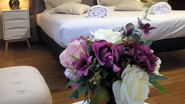 Une chambre à coucher avec un grand lit, deux tables de chevet avec des lampes, une table de nuit et un arrangement floral posé sur le sol.