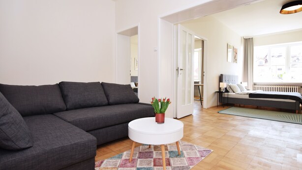 Ein modernes Wohnzimmer mit einem Ecksofa, einem runden weißen Couchtisch mit Topfpflanze und Blick auf ein Schlafzimmer mit Bett und Fenster.