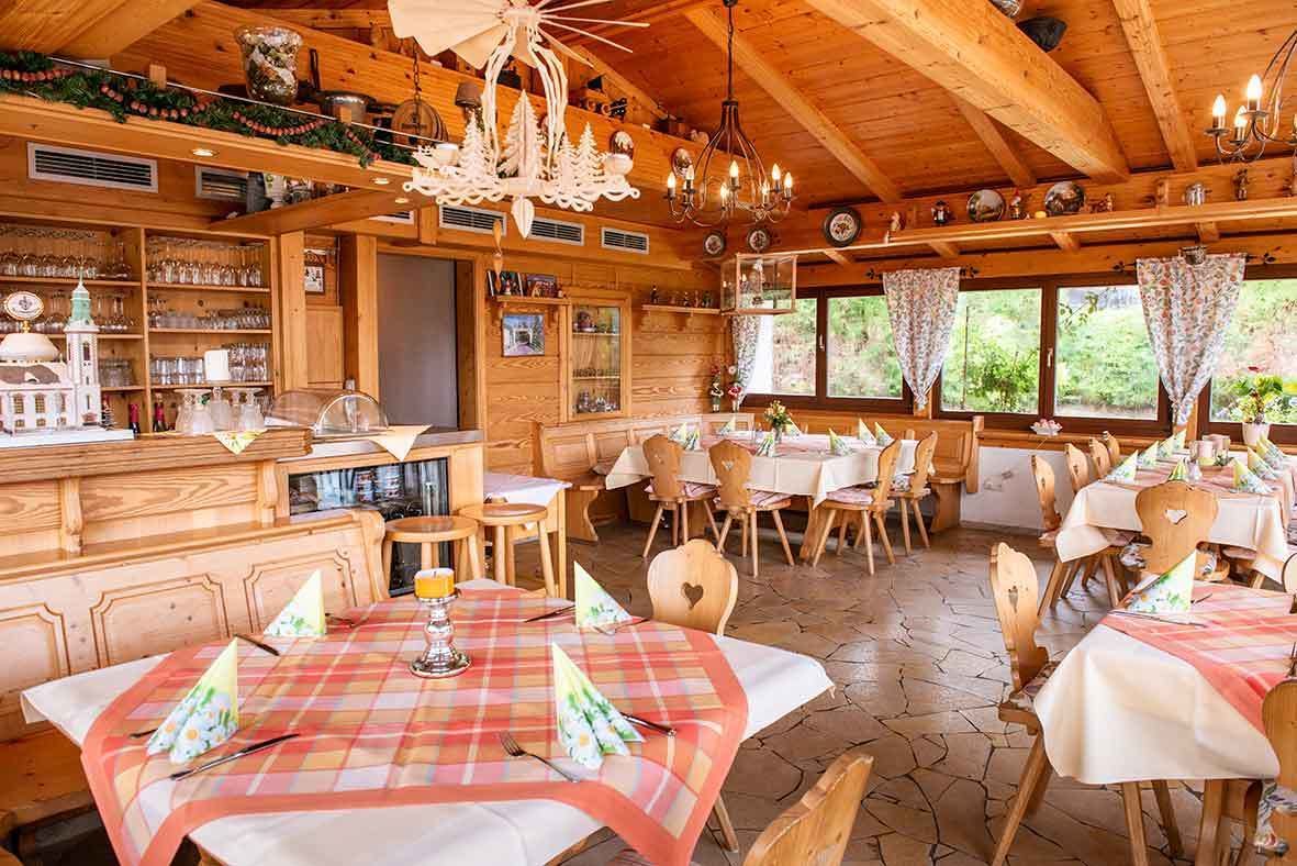 Interior rústico de madeira com várias mesas de jantar dispostas para hóspedes, um bar e vista para o verde lá fora.