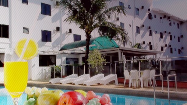 Zona de piscina con sillones de descanso, una mesa con fruta y una bebida, y un edificio con varias ventanas y balcones al fondo.
