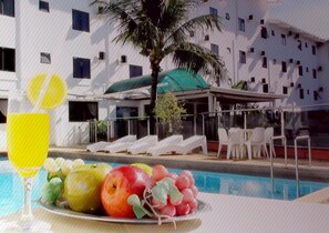 A poolside area with lounge chairs, a table with fruit and a drink, and a building with multiple windows and balconies in the background.