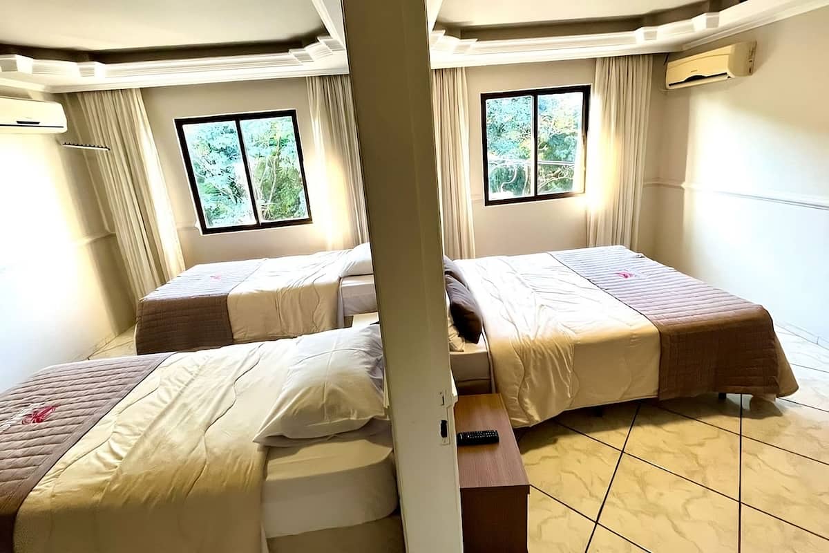 A hotel room with two beds, a window with a view of greenery, and an air conditioning unit on the wall.