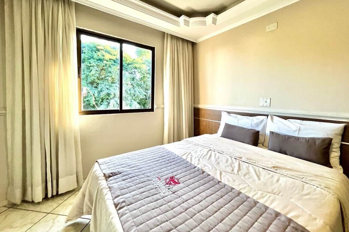 A hotel room with a bed, curtains, a window, and a wall outlet.