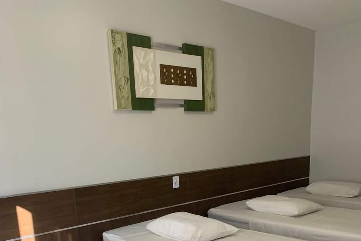 A hotel room with three single beds, each with white bedding and pillows. The room has a wooden headboard and a wall-mounted light fixture.