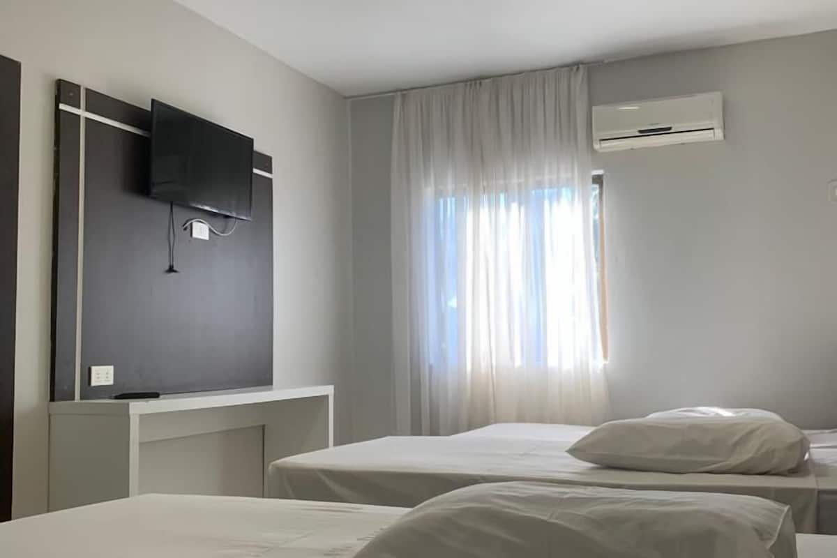 A hotel room with two single beds, a flat-screen TV mounted on the wall, an air conditioning unit, and a window with sheer curtains.