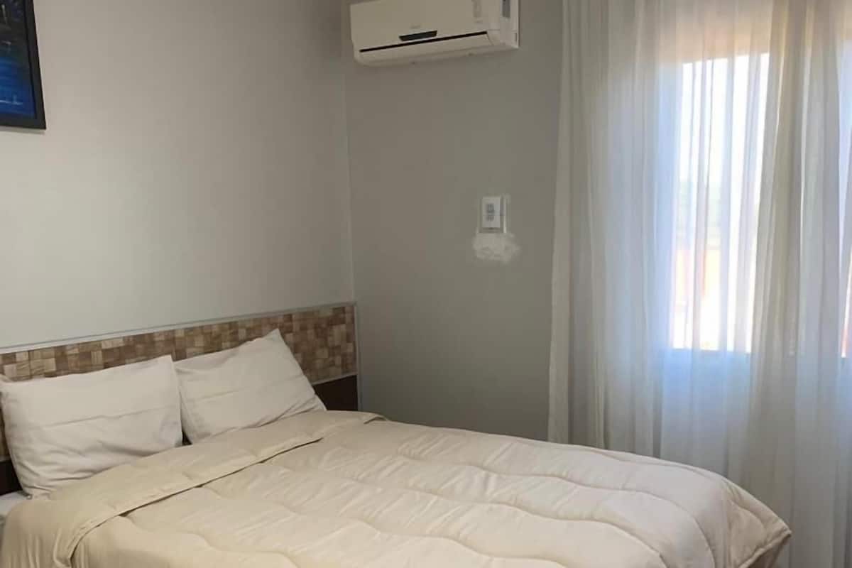 A hotel room with a bed, an air conditioning unit, a window with curtains, and a tiled floor.