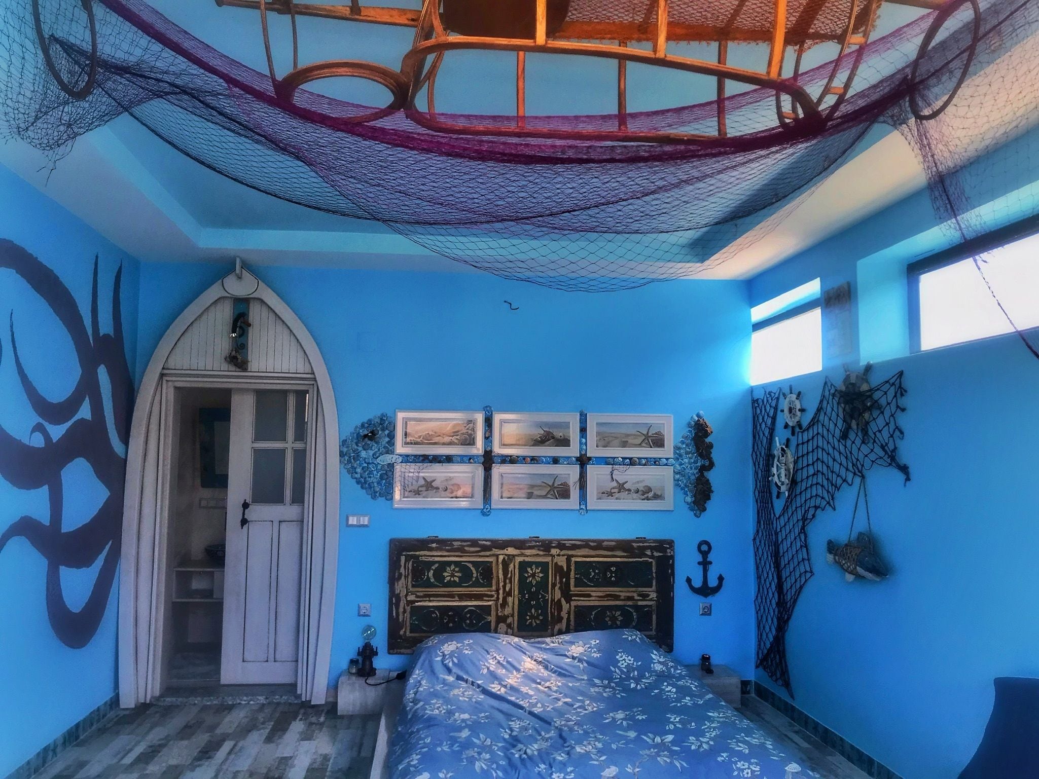 Una habitación con pared azul, cama con colcha floral, cabecera de madera y hamaca en el techo.