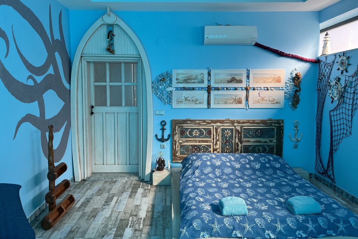 A bedroom with a nautical theme, featuring a bed with a blue and white pattern, a white door with a window, and wall decorations including framed pictures and a ship's wheel.