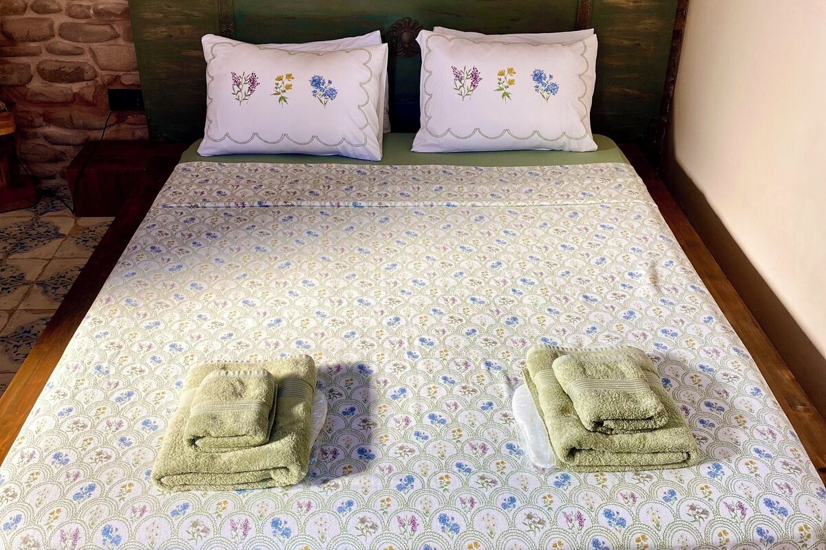 A bed with floral-patterned bedding, two decorative pillows with embroidered flowers, and two folded towels on the bed.
