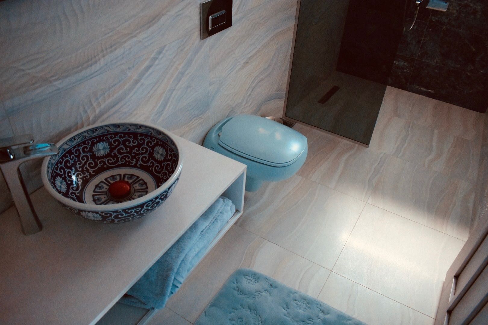 Une salle de bain avec un lavabo blanc, un bol décoratif, des toilettes bleues et une serviette posée sur le comptoir.