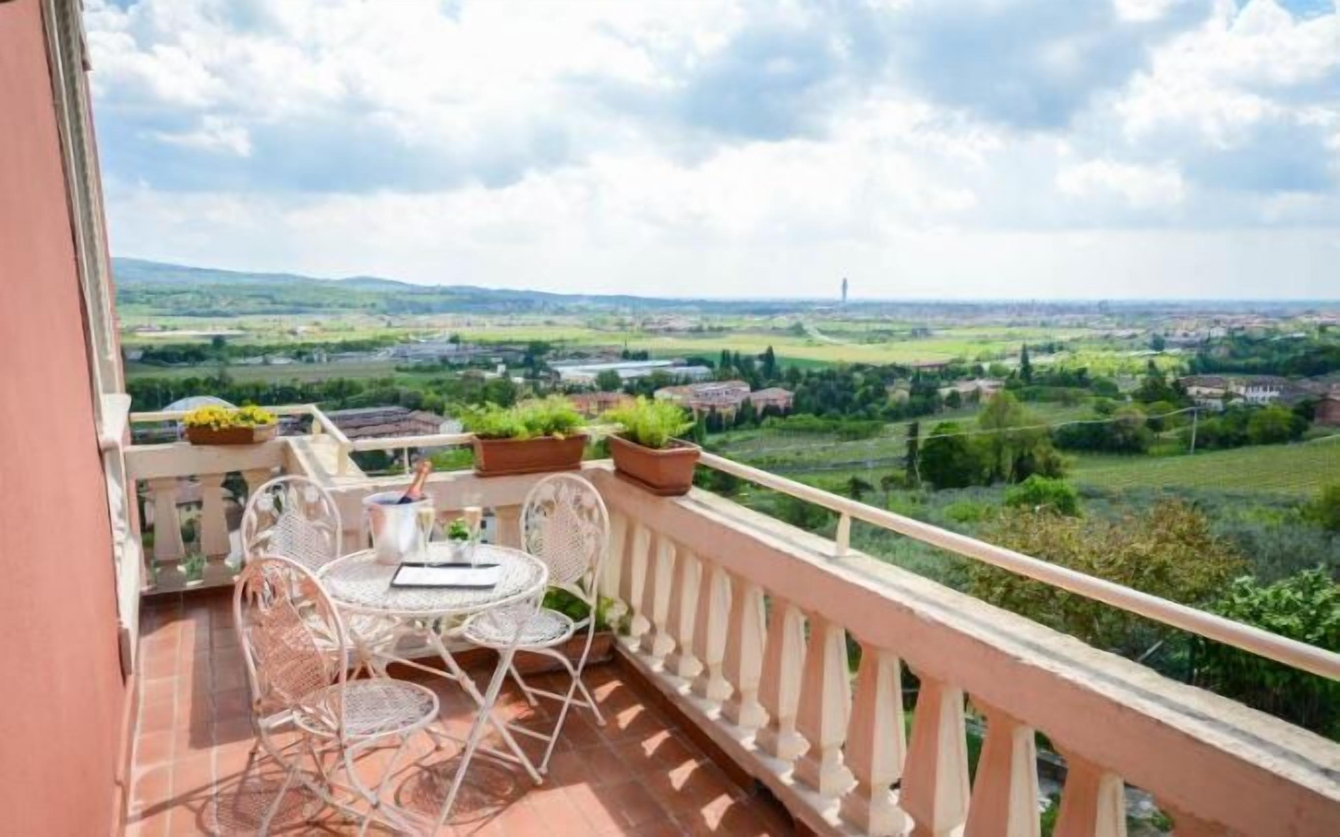 A balcony with a table and chairs, potted plants, and a view of a rural landscape.