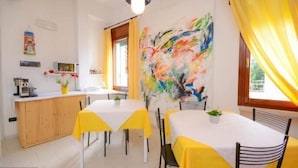 Ein Essbereich mit zwei Tischen, einer mit gelber Tischdecke und der andere mit weißer Tischdecke. An der Wand hängt ein farbenfrohes abstraktes Gemälde, ein Holzschrank und auf der Arbeitsplatte steht eine Kaffeemaschine.