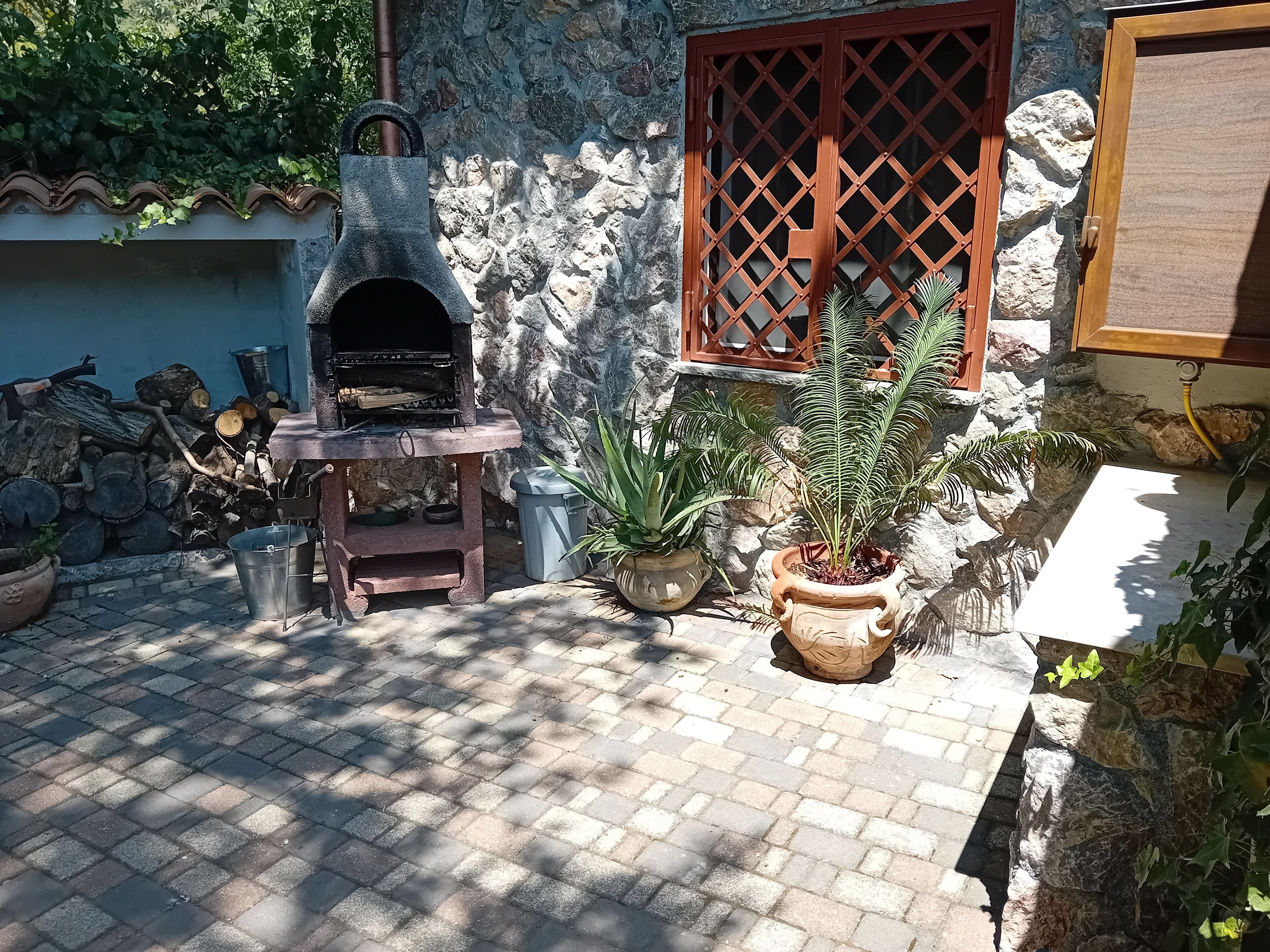 An outdoor patio with a stone wall, a wooden table, a barbecue grill, and potted plants.